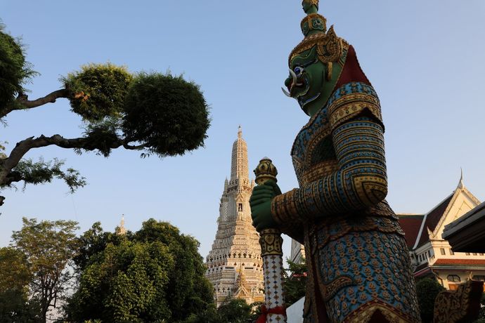 Op de voorgrond staat een groen, sierlijk, gigantisch standbeeld, met op de achtergrond de hoge, ingewikkelde witte torenspits van Wat Arun.