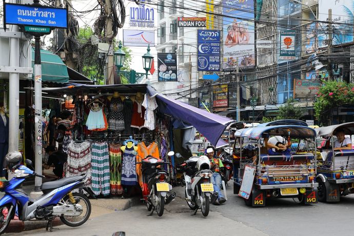 Een bruisend straatbeeld in Bangkok met kledingstalletjes, geparkeerde motoren en tuk-tuks onder een wirwar van bovengrondse elektriciteitskabels.