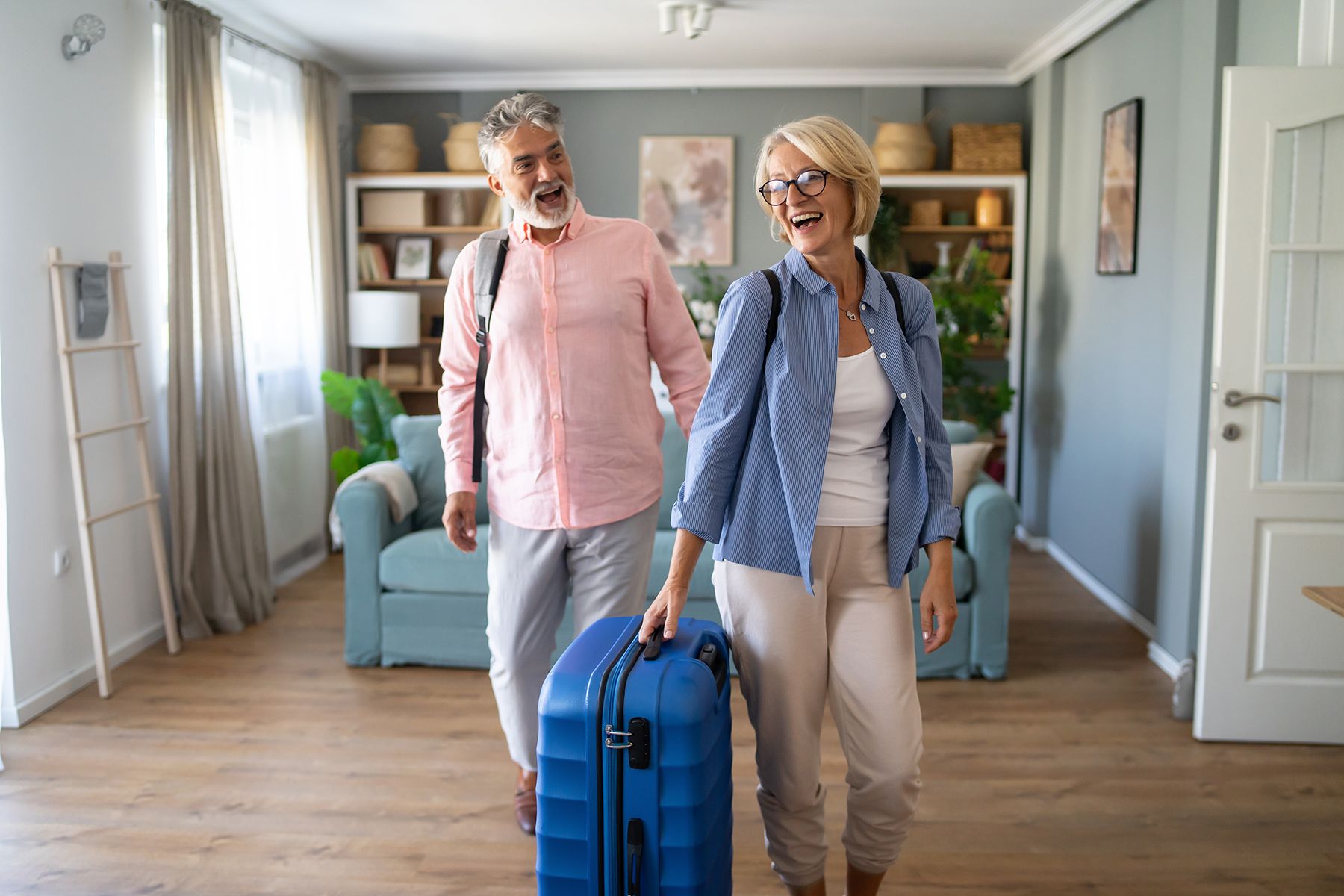 An elderly couple is walking in a living room with their luggage.