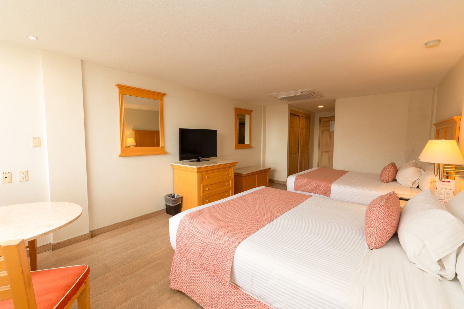 Habitación de hotel con dos camas dobles, TV, cómoda, lámpara y decoración en tonos beige.