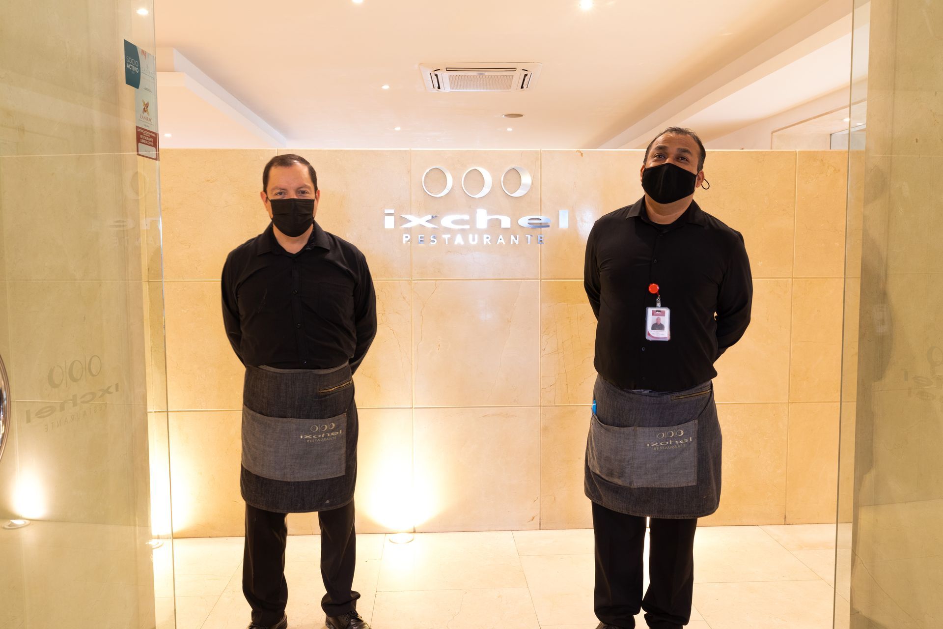 Dos empleados enmascarados permanecen de pie en un pasillo iluminado, vestidos con uniformes negros y delantales grises.