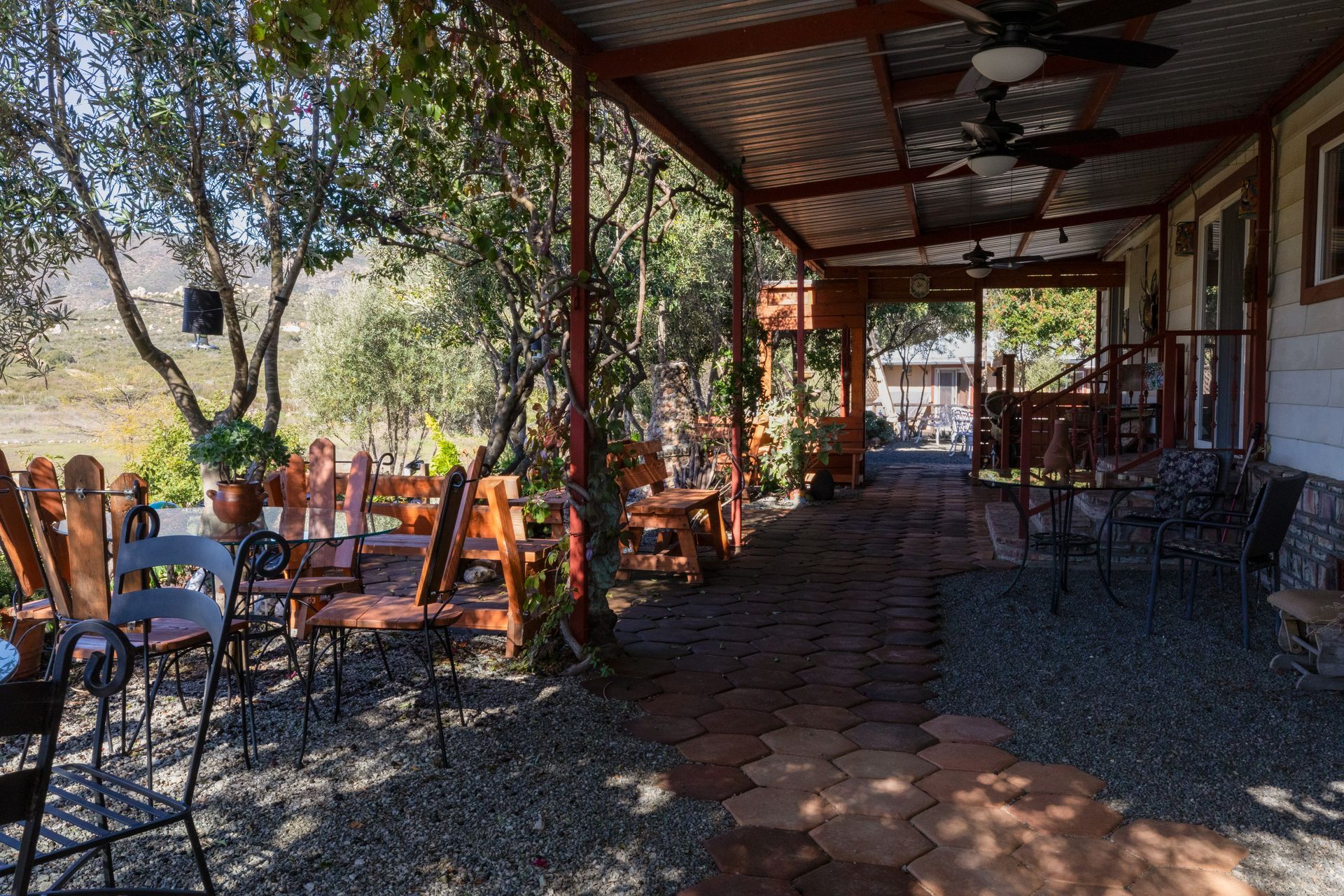 Terraza sombreada al aire libre con mesas, sillas y una pérgola de techo rojo entre árboles y un sendero de grava.