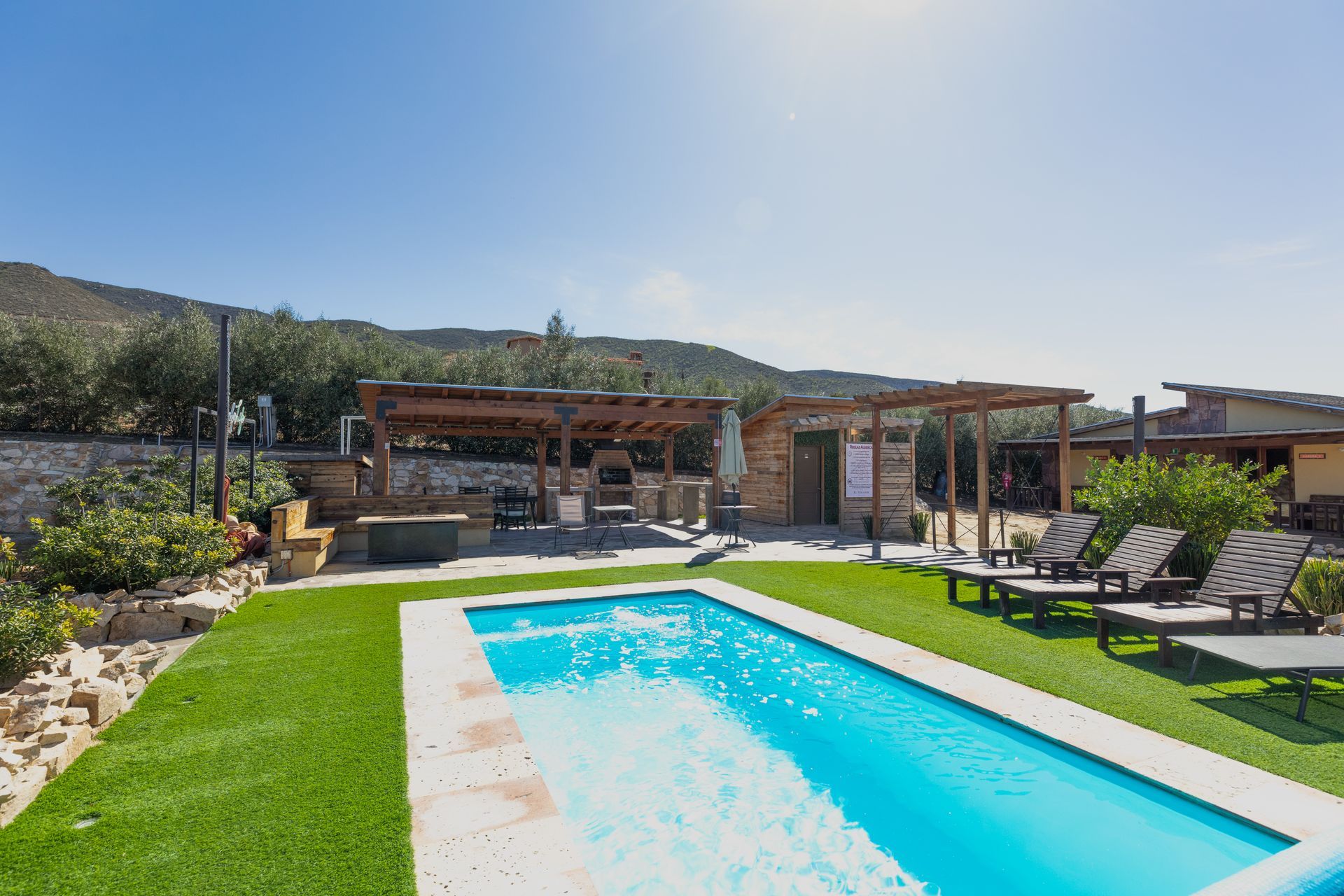 Jardín moderno con piscina turquesa, tumbonas y patio sombreado con vistas a la montaña.