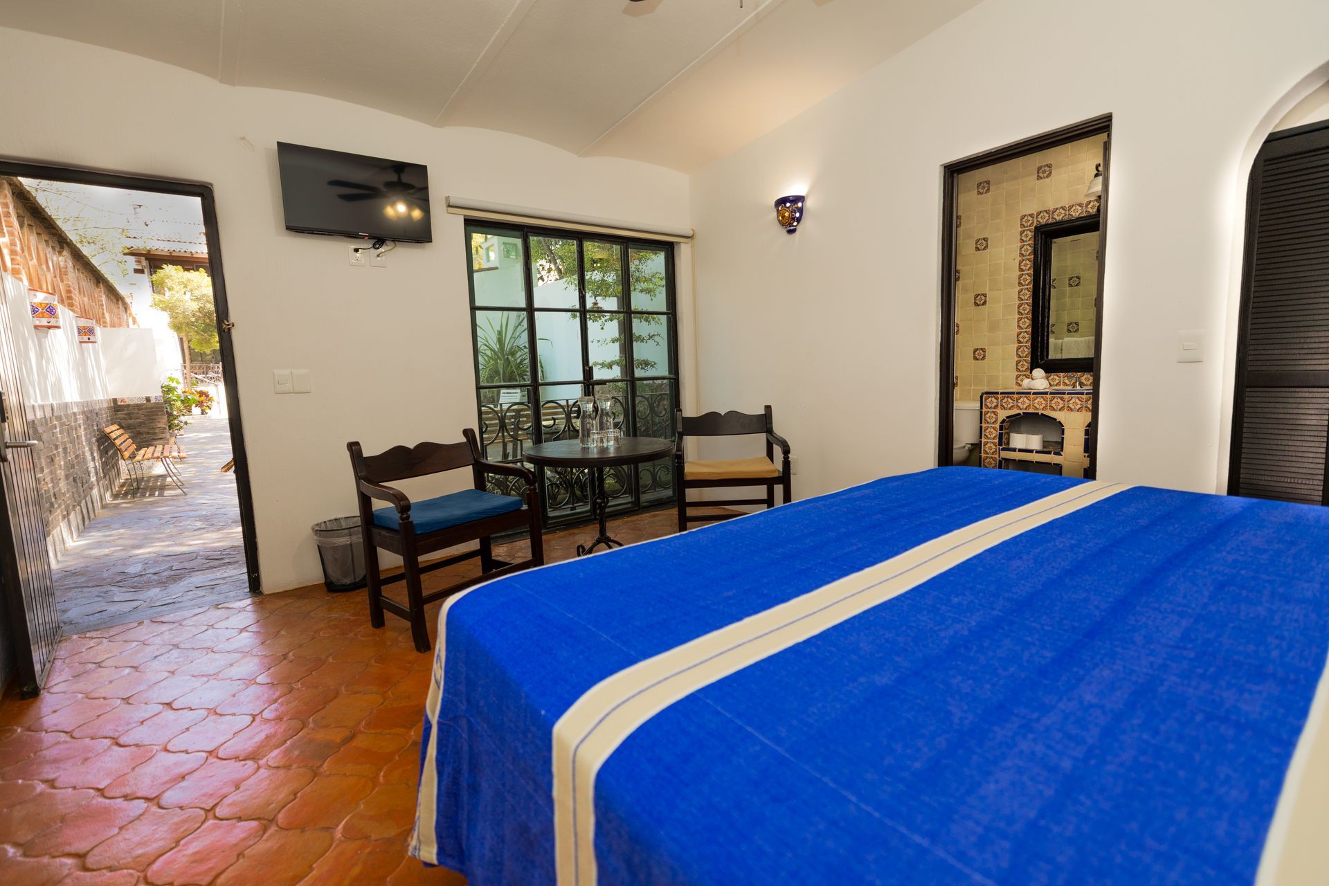 Habitación de hotel luminosa con colcha azul, suelo de baldosas de terracota y vistas al patio a través de las puertas abiertas.