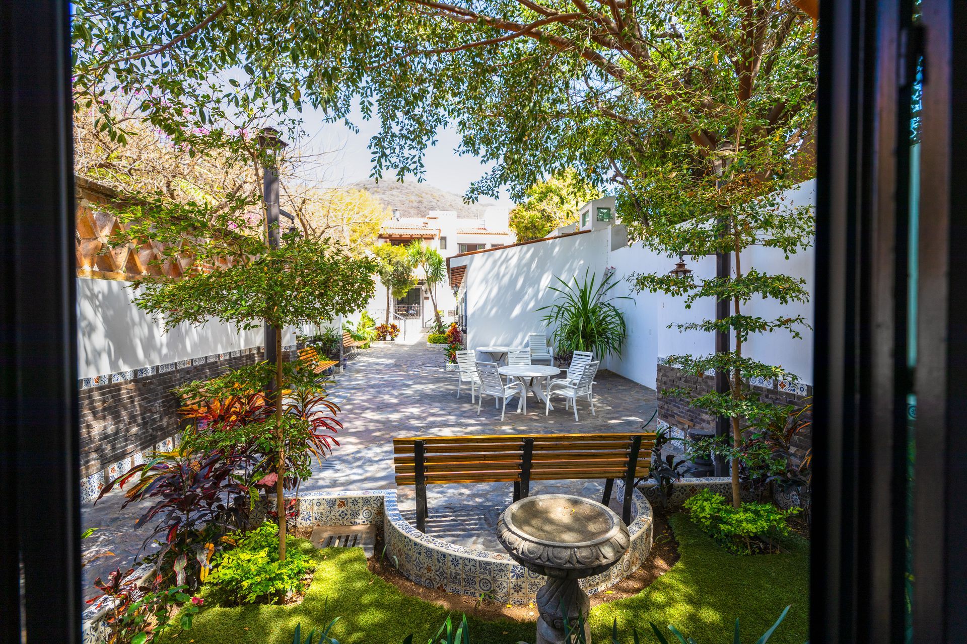 Patio sombreado con jardín, banco de madera, mesa de piedra y sillas blancas junto a un sendero soleado.