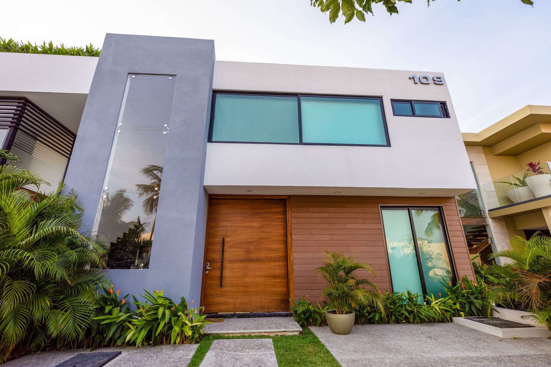 Casa moderna de dos plantas con fachada gris y blanca, puertas de entrada de madera, grandes ventanales azules y un exuberante jardín.