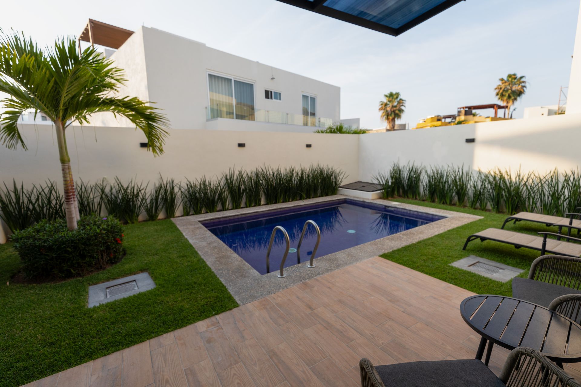 Patio trasero moderno con una pequeña piscina azul, tumbonas y una casa blanca bajo un patio cubierto.