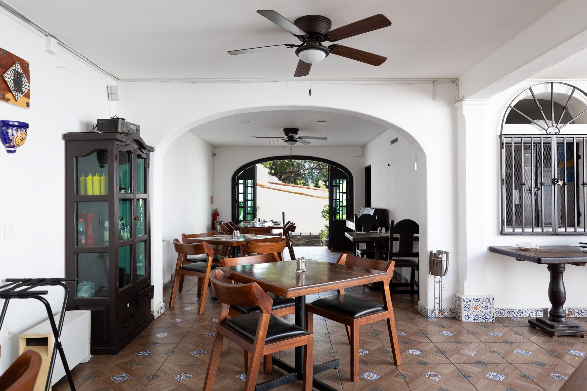 Comedor interior con mesas y sillas de madera, ventilador de techo, puerta arqueada y suelo de baldosas.