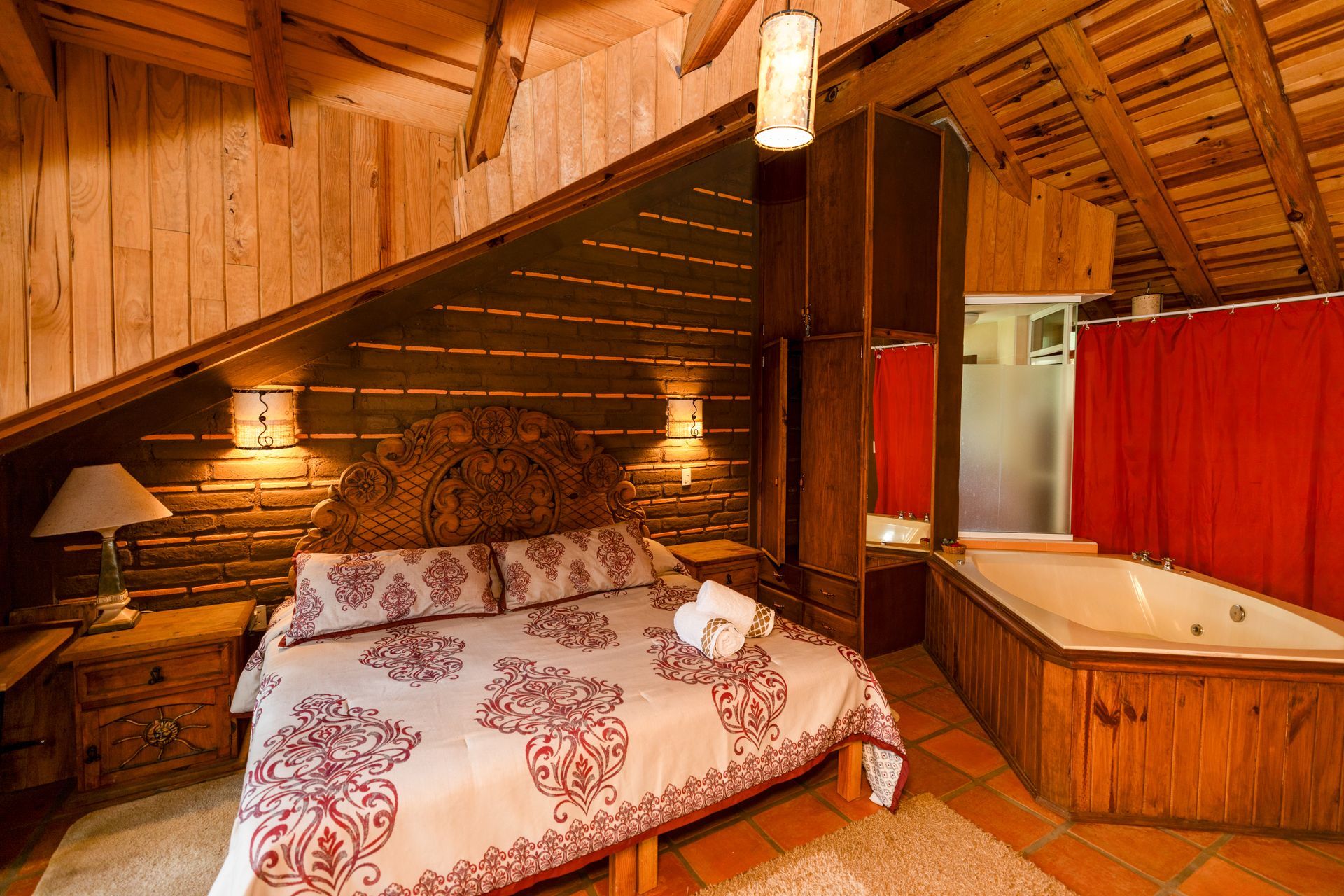 Acogedor dormitorio abuhardillado con cama, techo de madera inclinado, cortinas rojas e iluminación cálida.