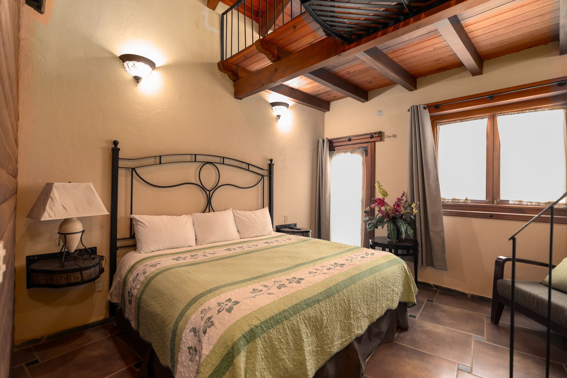 Acogedora habitación con cama de hierro forjado, edredón verde, vigas de madera en el techo y dos ventanas.