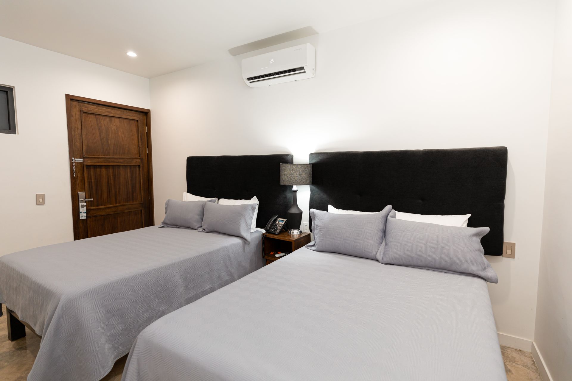 Habitación bien decorada con dos camas grises, cabeceros negros, puerta de madera y aire acondicionado mural.