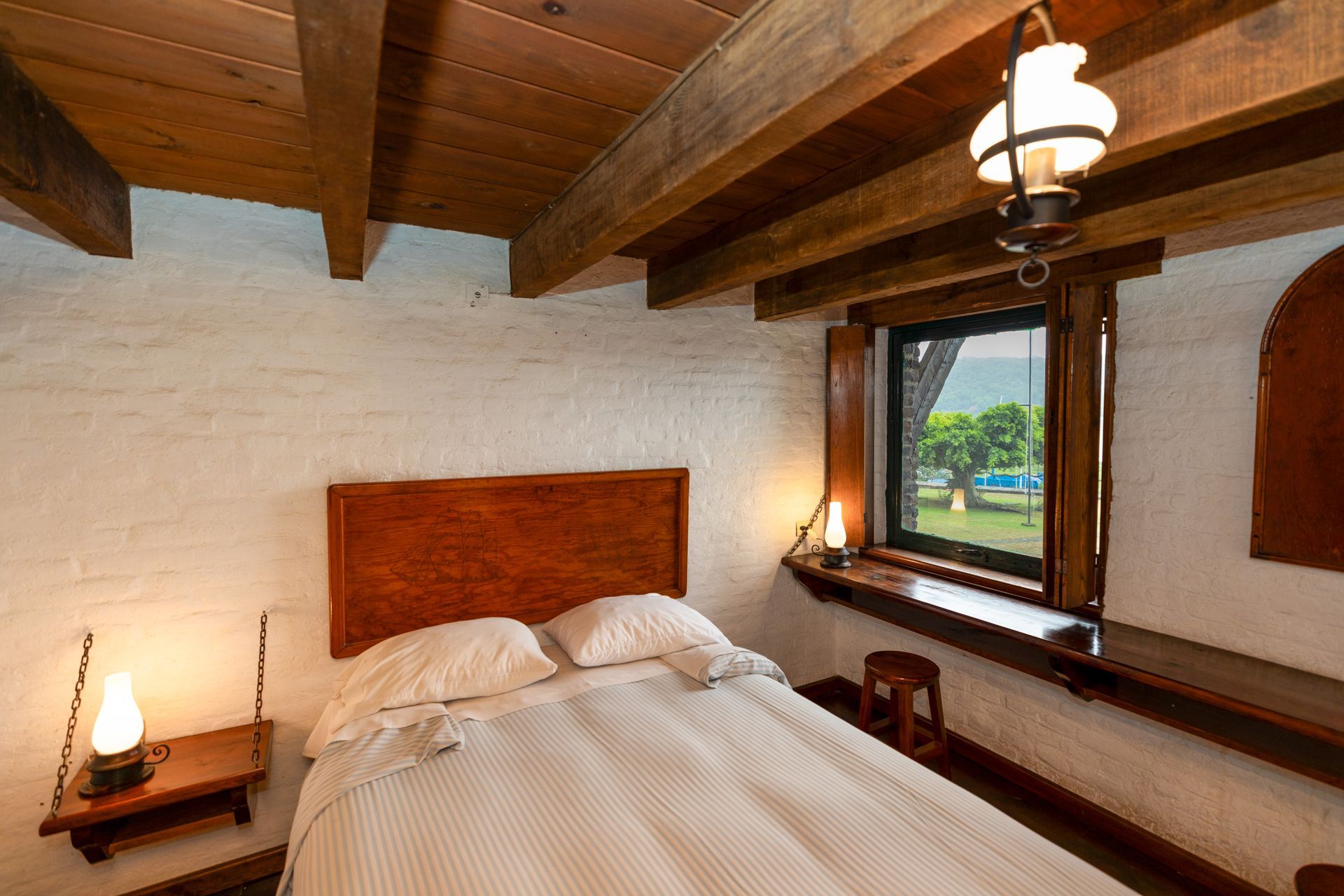 Acogedora habitación con vigas de madera en el techo, paredes de ladrillo blanco y vistas a la vegetación desde la ventana.