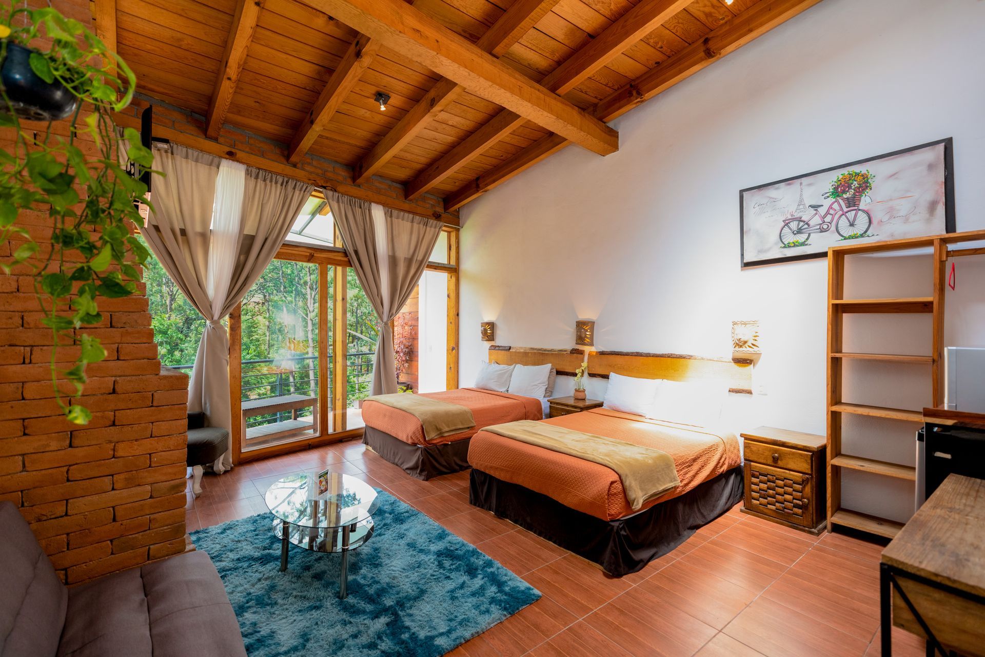 Habitación acogedora con techo de madera, grandes ventanales, colcha naranja, alfombra azul y silla de mimbre.