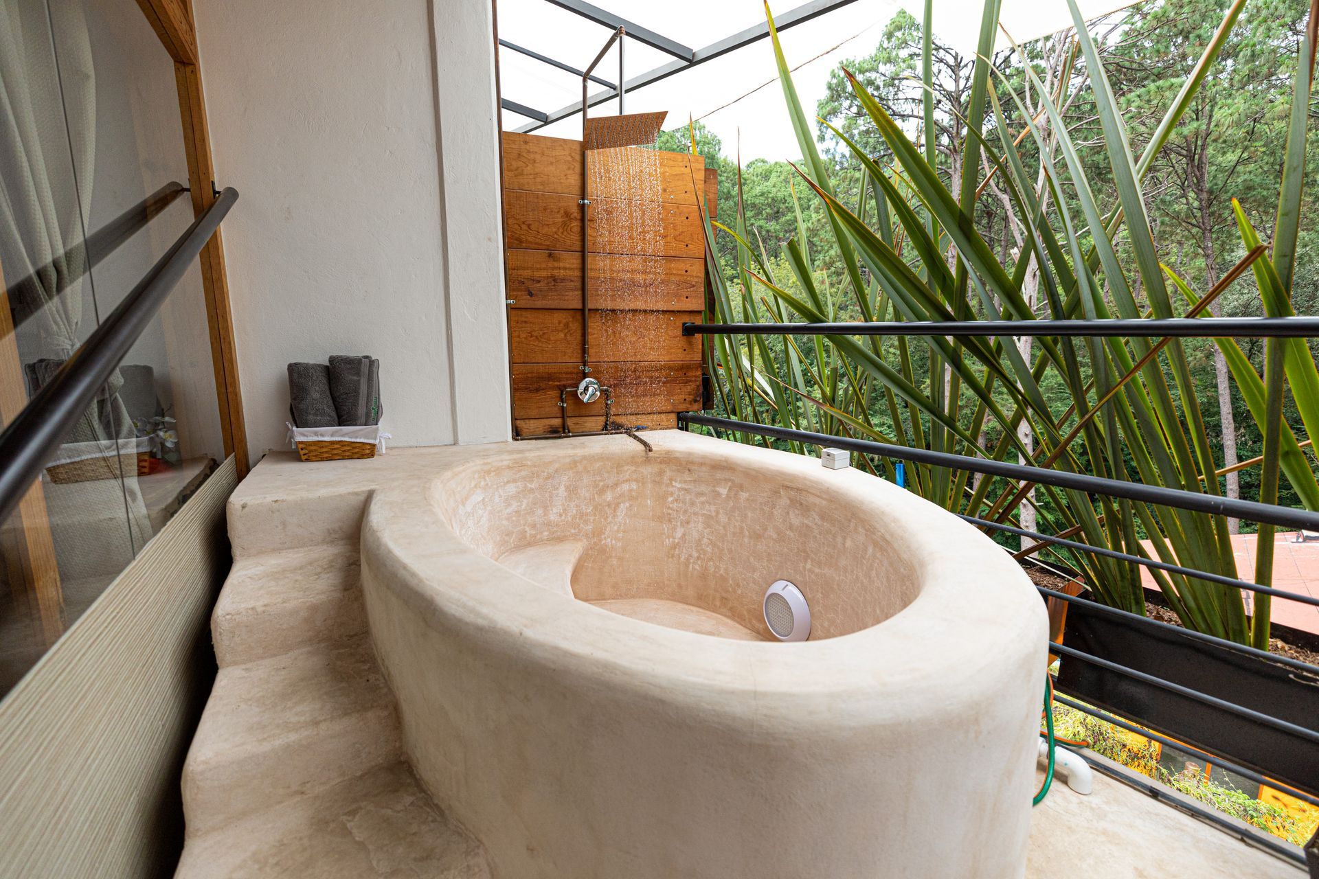 Baño exterior con bañera y lavabo de piedra beige en un balcón, rodeado de vegetación.