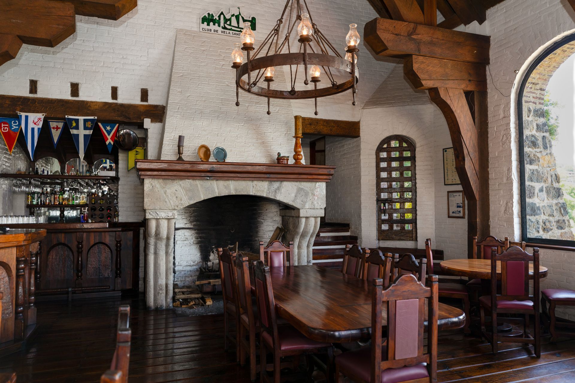 Interior rústico de pub con mesas de madera, chimenea de piedra, ventanas arqueadas y una lámpara de araña colgante.
