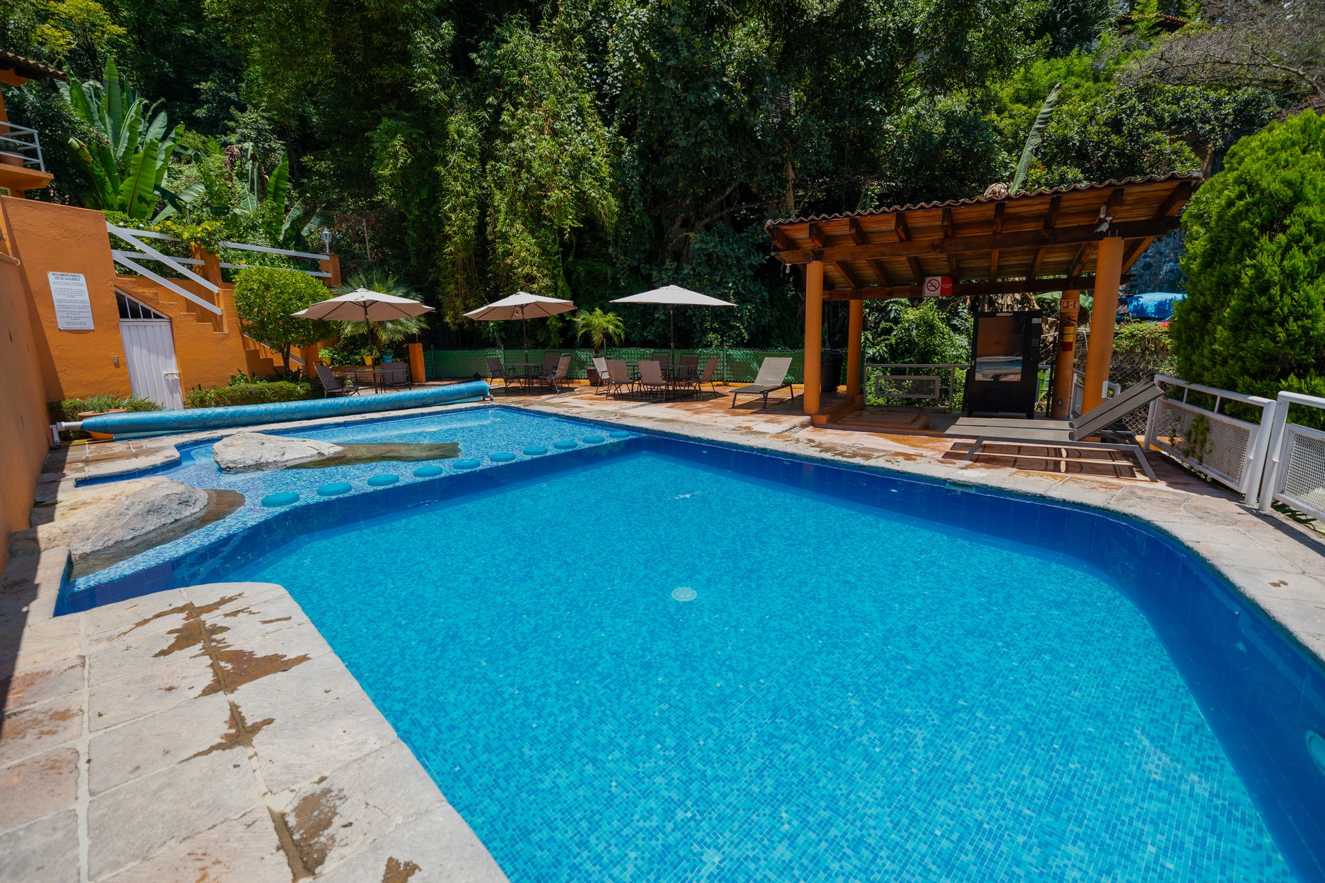 Piscina al aire libre con agua azul brillante, terraza de piedra, árboles, tumbonas y pérgola con sombra.