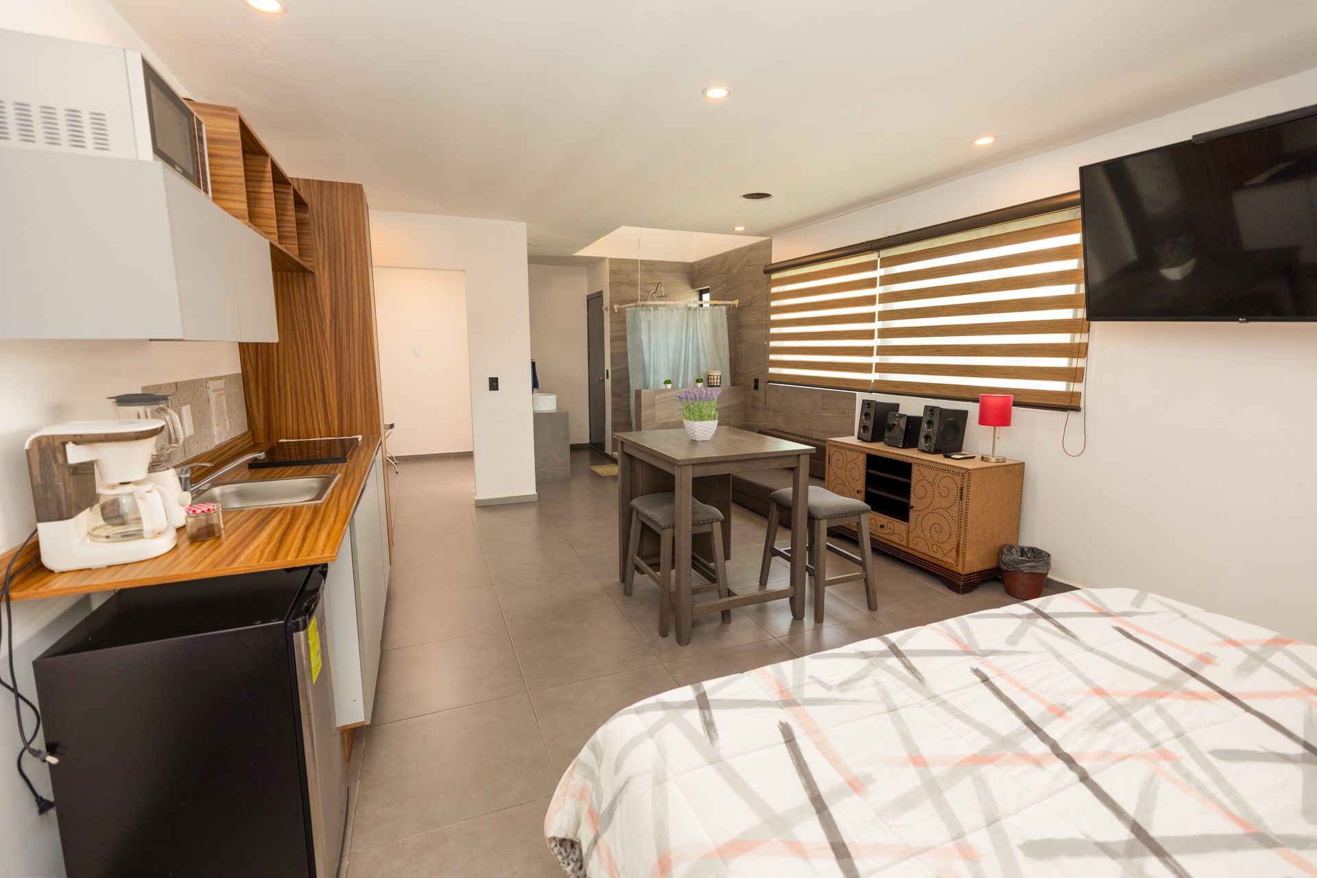 Apartamento tipo estudio moderno con cocina, mesa de comedor, cama, televisor y persianas a rayas.