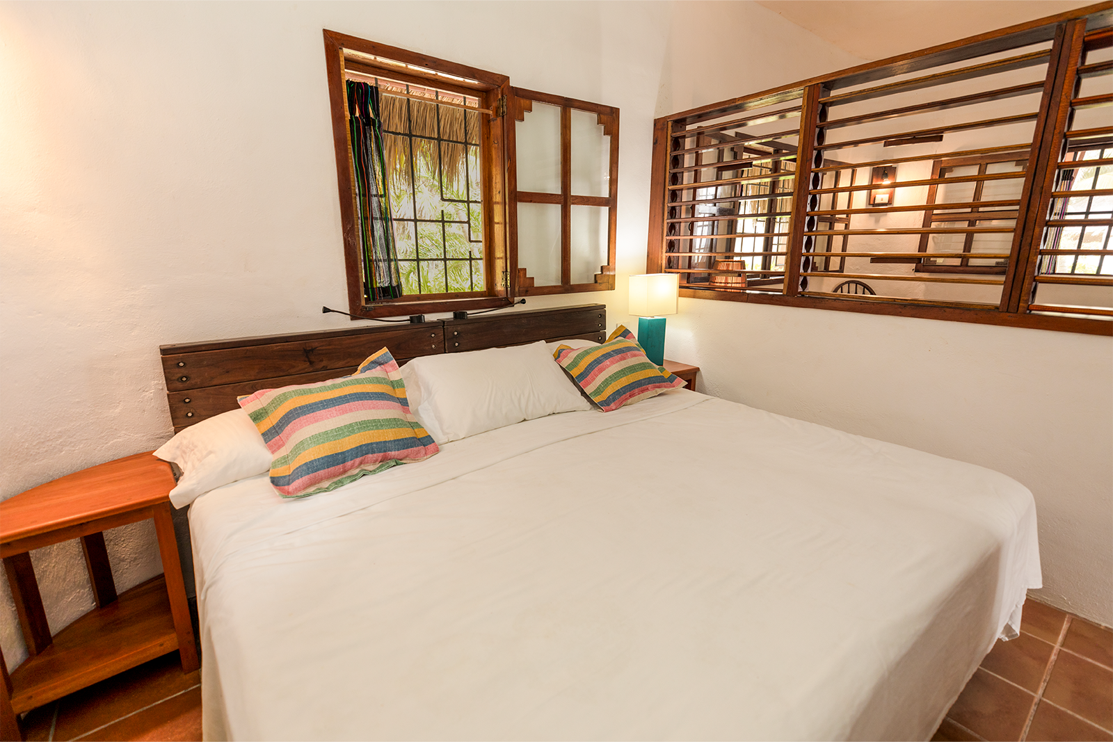 Habitación luminosa con una cama grande blanca, almohadas a rayas, ventanas de madera y suelo de baldosas de terracota.