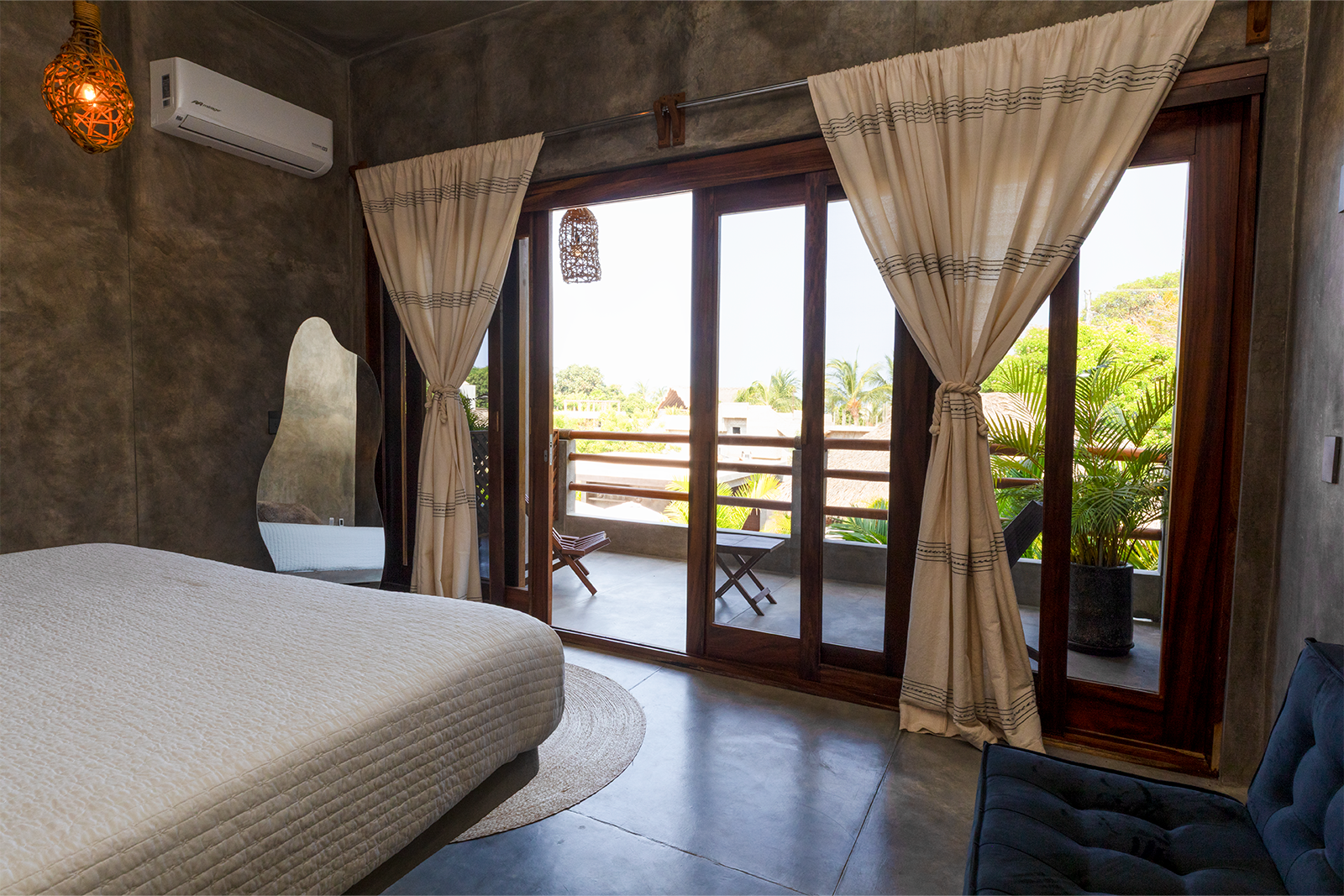 Habitación luminosa con cama, cortinas, puertas que dan al balcón y vistas tropicales soleadas.