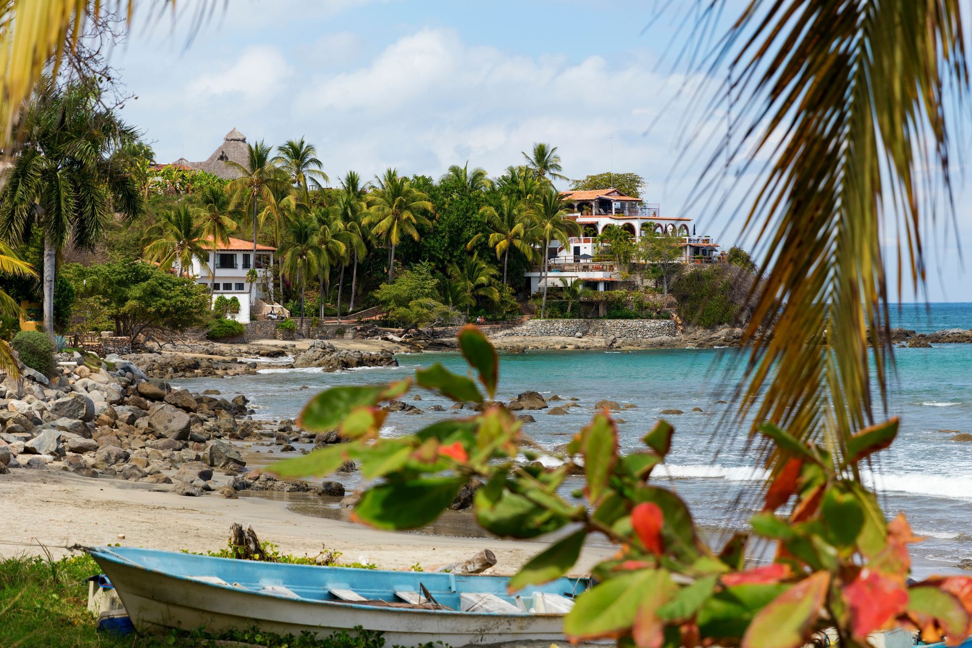 Cala tropical con una pequeña embarcación, costa rocosa, palmeras y casas junto al mar.