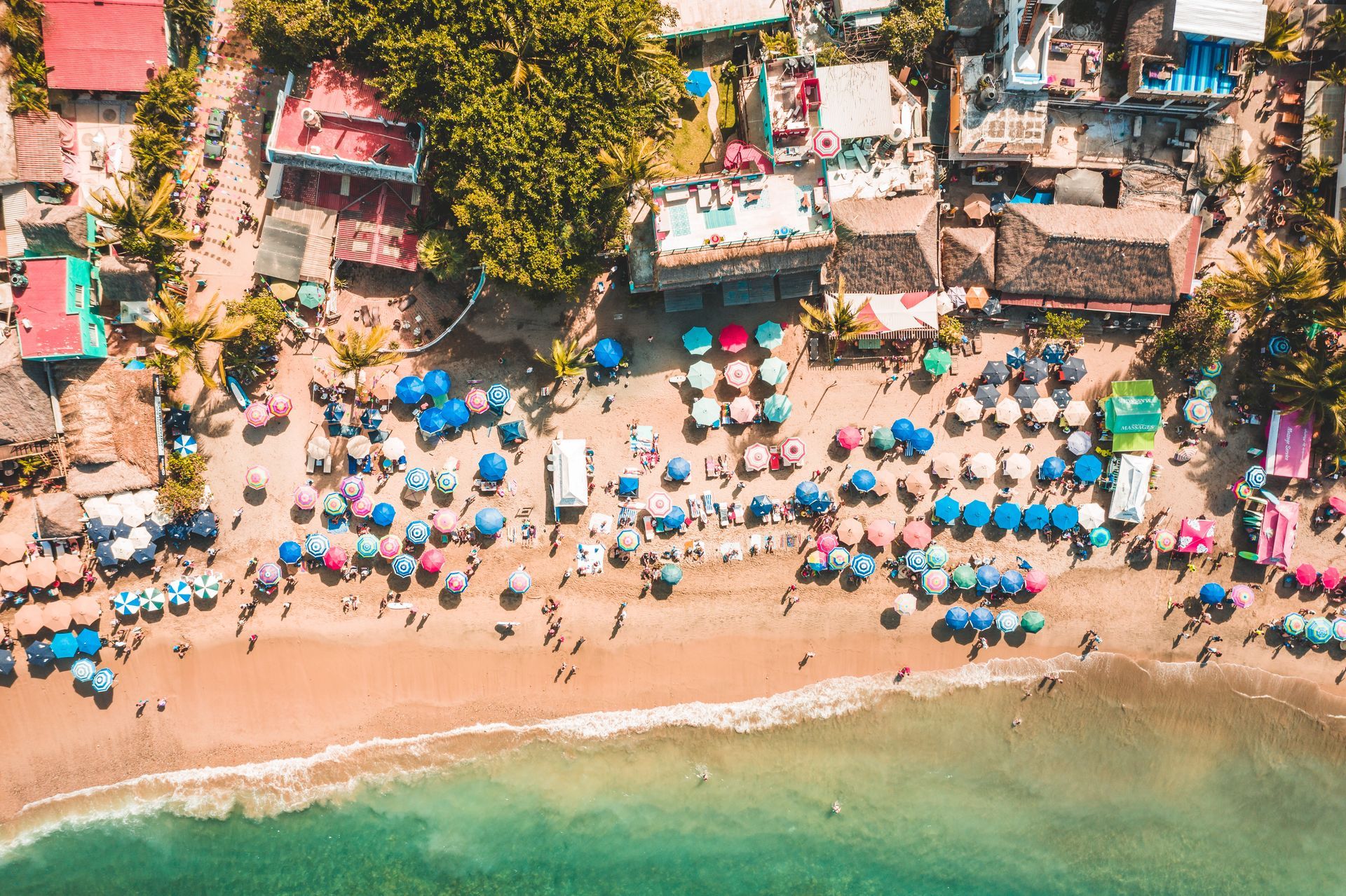 Vista aérea de una playa abarrotada con sombrillas azules, la costa y agua turquesa junto a cabañas costeras.