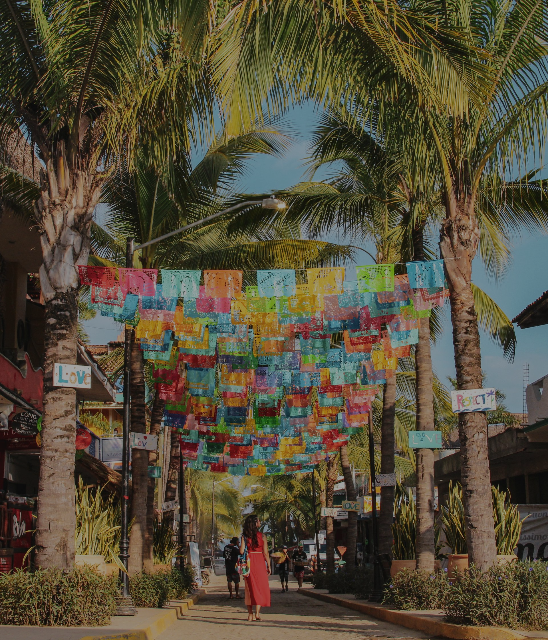 Un paseo bordeado de palmeras con coloridas pancartas colgantes y una persona con un vestido rojo de pie abajo.