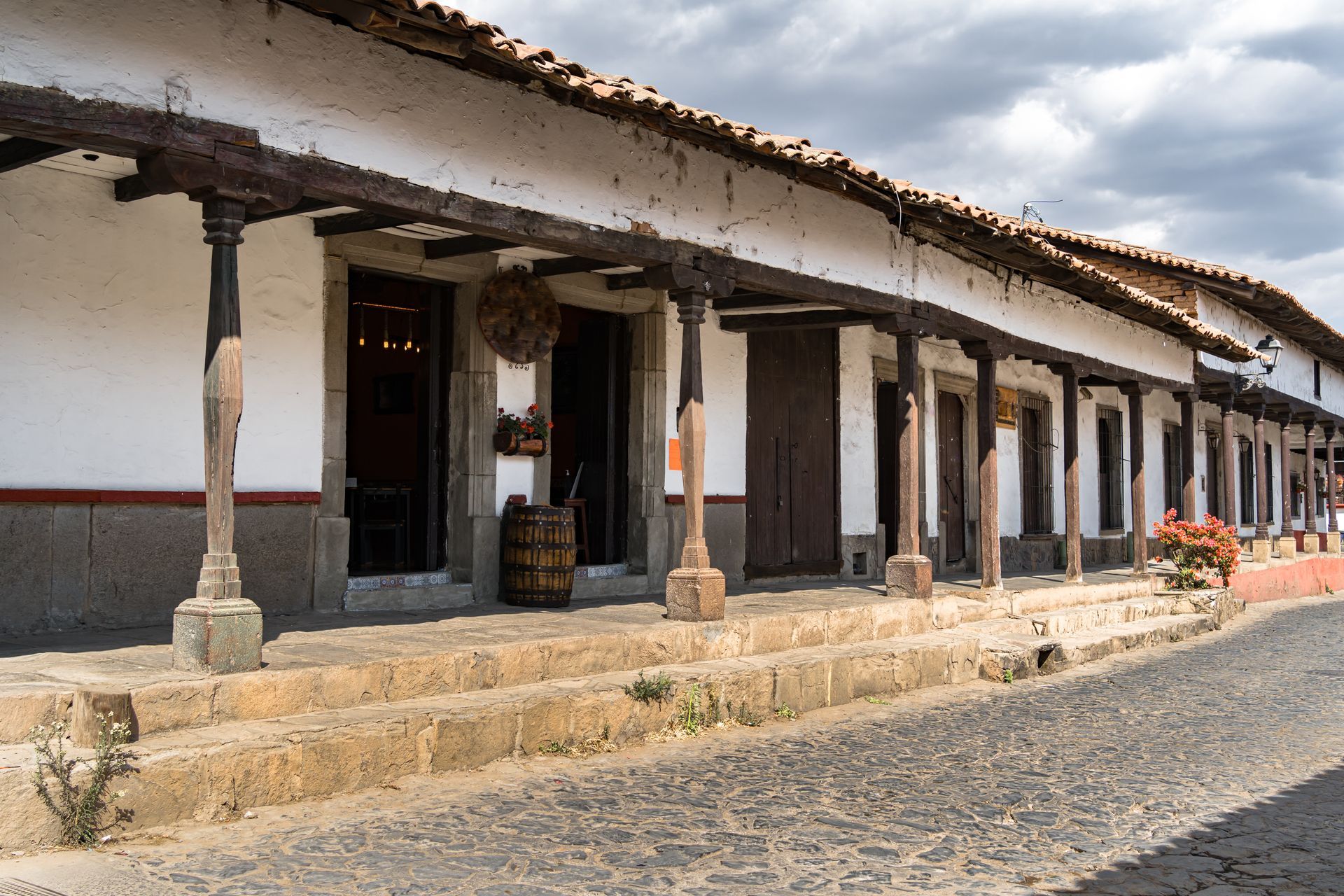 Edificio tradicional de adobe con columnas de madera y un camino de piedra en una calle soleada.