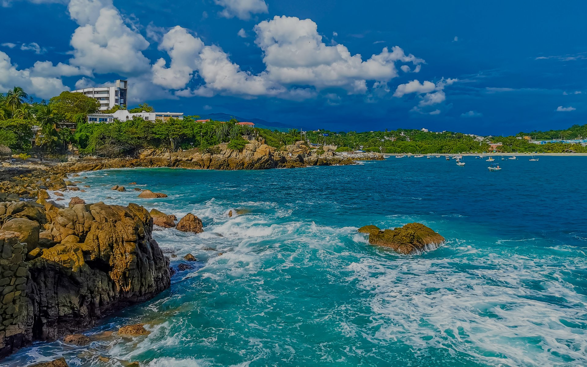 Costa tropical rocosa con olas turquesas, un cielo azul y edificios distantes bajo nubes blancas.