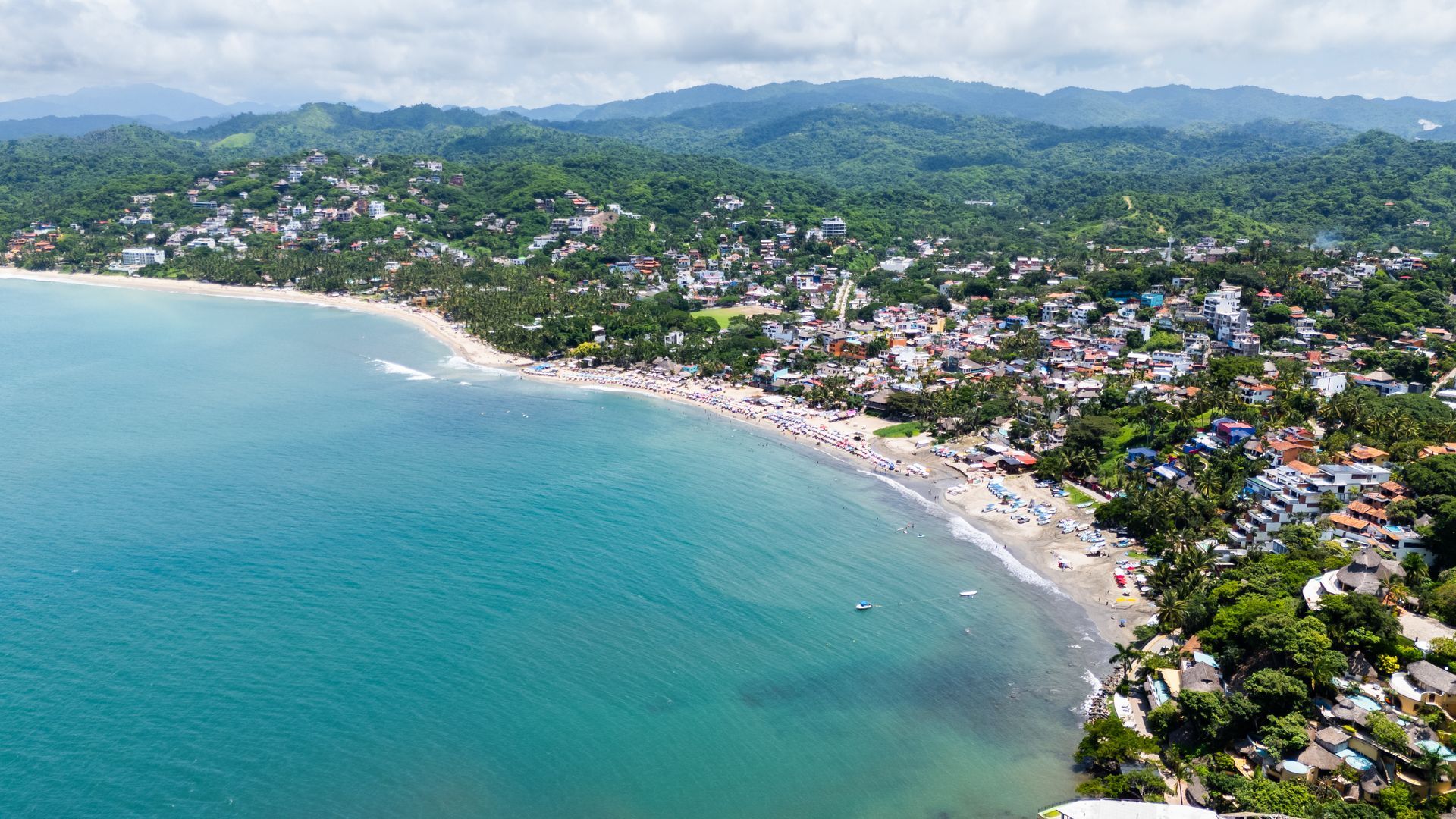 Vista aérea de una bahía turquesa, una playa de arena y un pueblo costero densamente poblado de vegetación, con colinas al fondo.