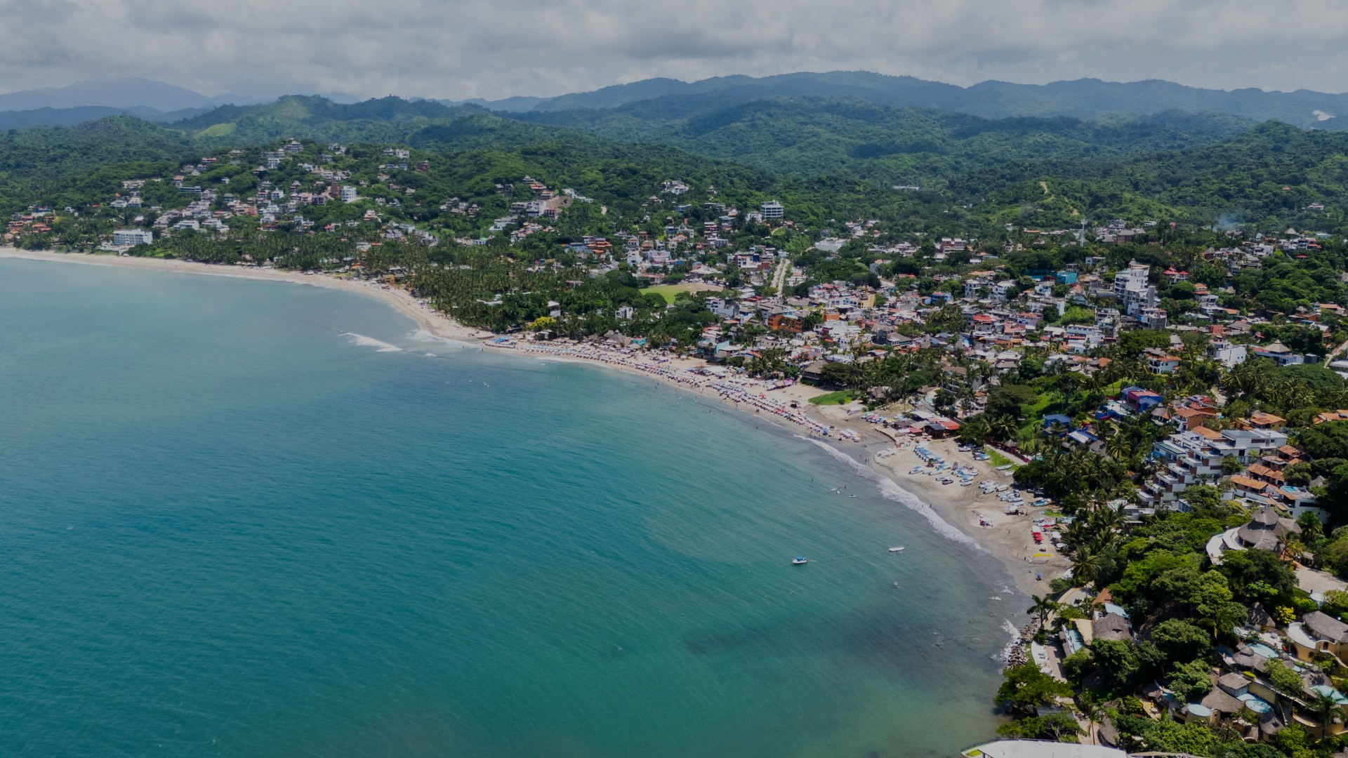 Vista aérea de una playa en forma de media luna con aguas turquesas, un pueblo costero y colinas verdes al fondo.