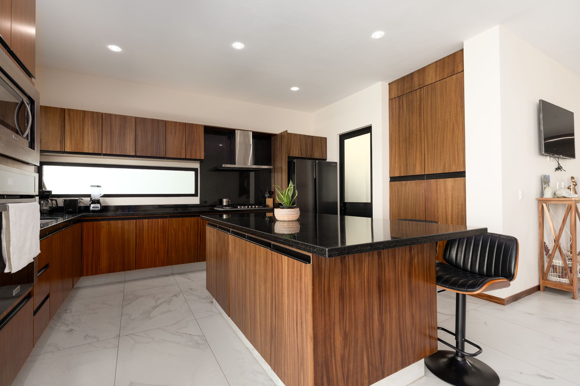 Cocina moderna con gabinetes de madera oscura, piso de baldosas blancas, isla y taburetes de bar negros.