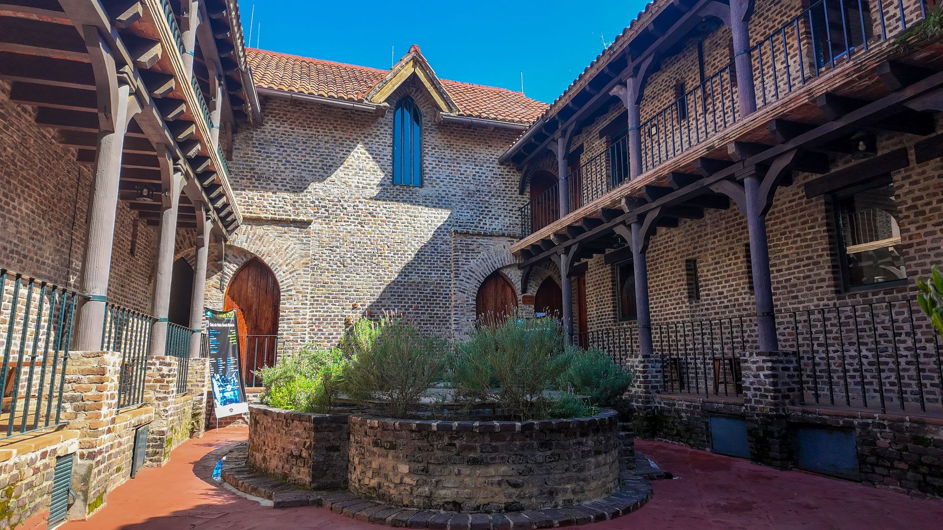 Patio de piedra con balcones de madera, puerta arqueada y una jardinera circular bañada por la cálida luz del sol.