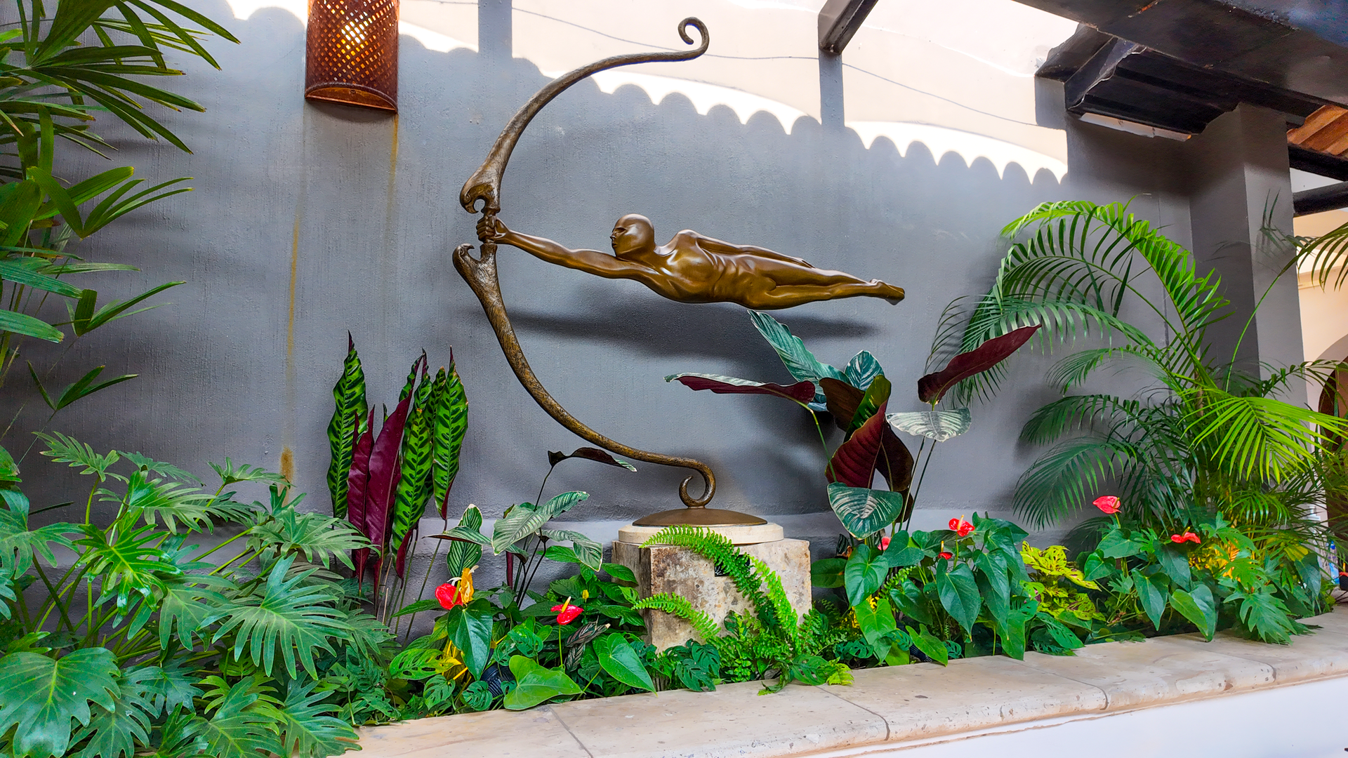Fuente de jardín con una figura voladora de bronce, exuberantes plantas verdes y flores rojas contra un muro de piedra.