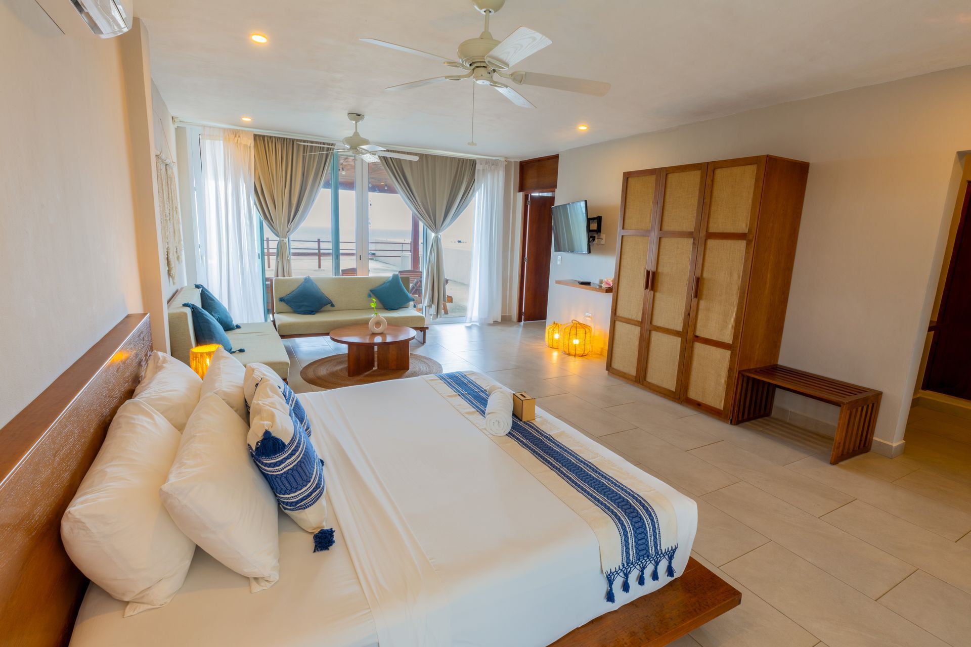 Habitación luminosa con cama blanca, armario de madera, ventilador de techo y puertas de cristal que dan a un balcón.