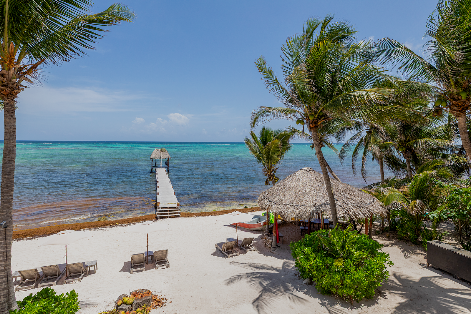 Playa tropical con arena blanca, agua turquesa, tumbonas, palmeras y una cabaña con techo de paja bajo el cielo azul.