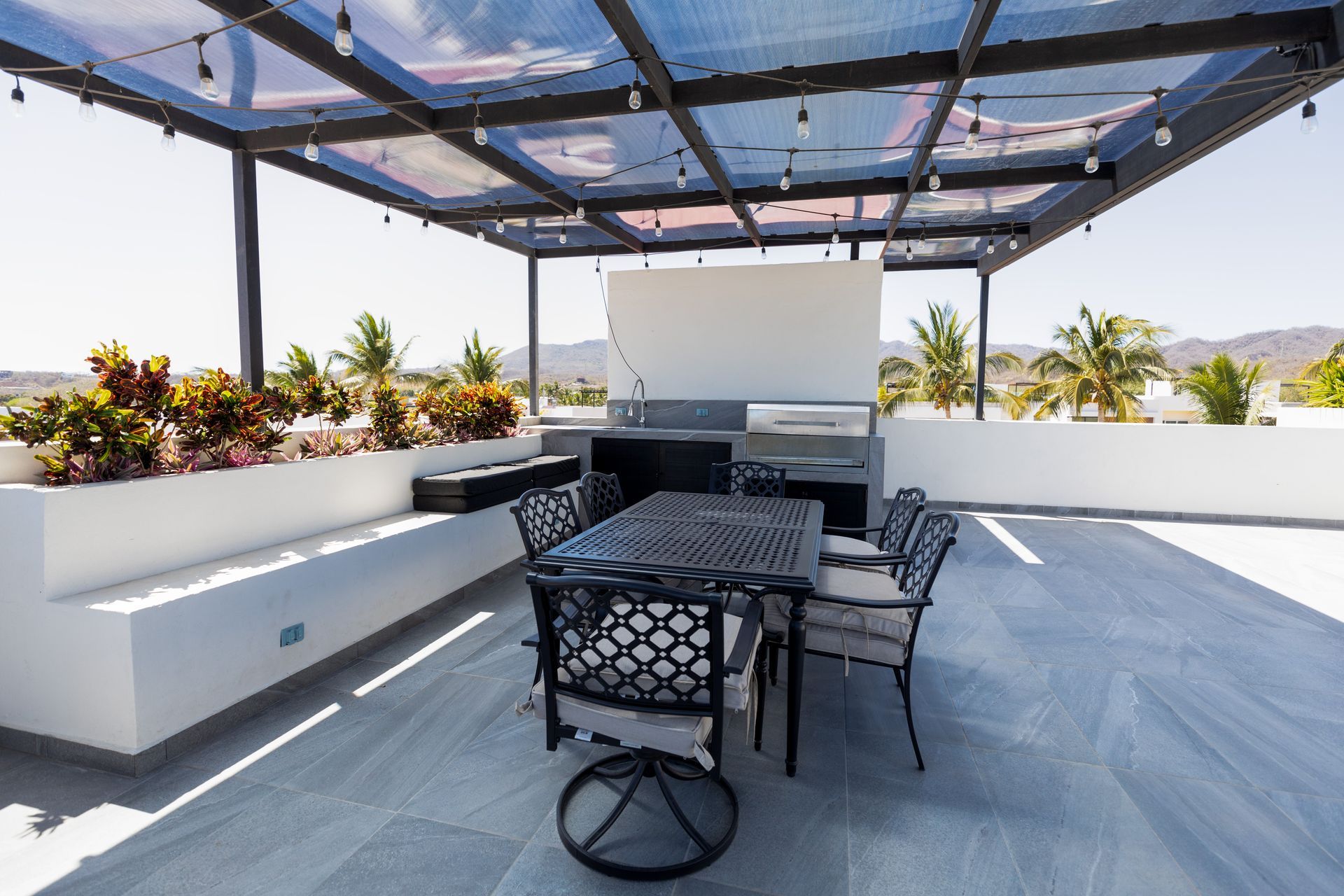Terraza moderna en la azotea con conjunto de comedor de metal negro, asientos empotrados y pérgola con vistas a las palmeras.