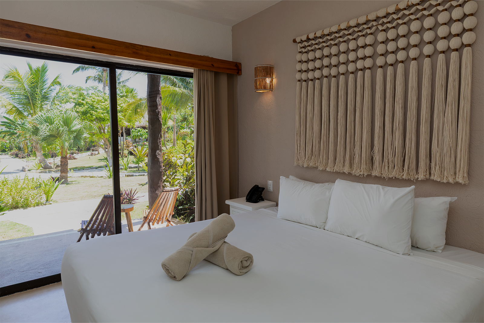 Habitación luminosa junto a la playa con cama blanca, decoración de pared de macramé y puertas correderas que dan a palmeras y a un patio.
