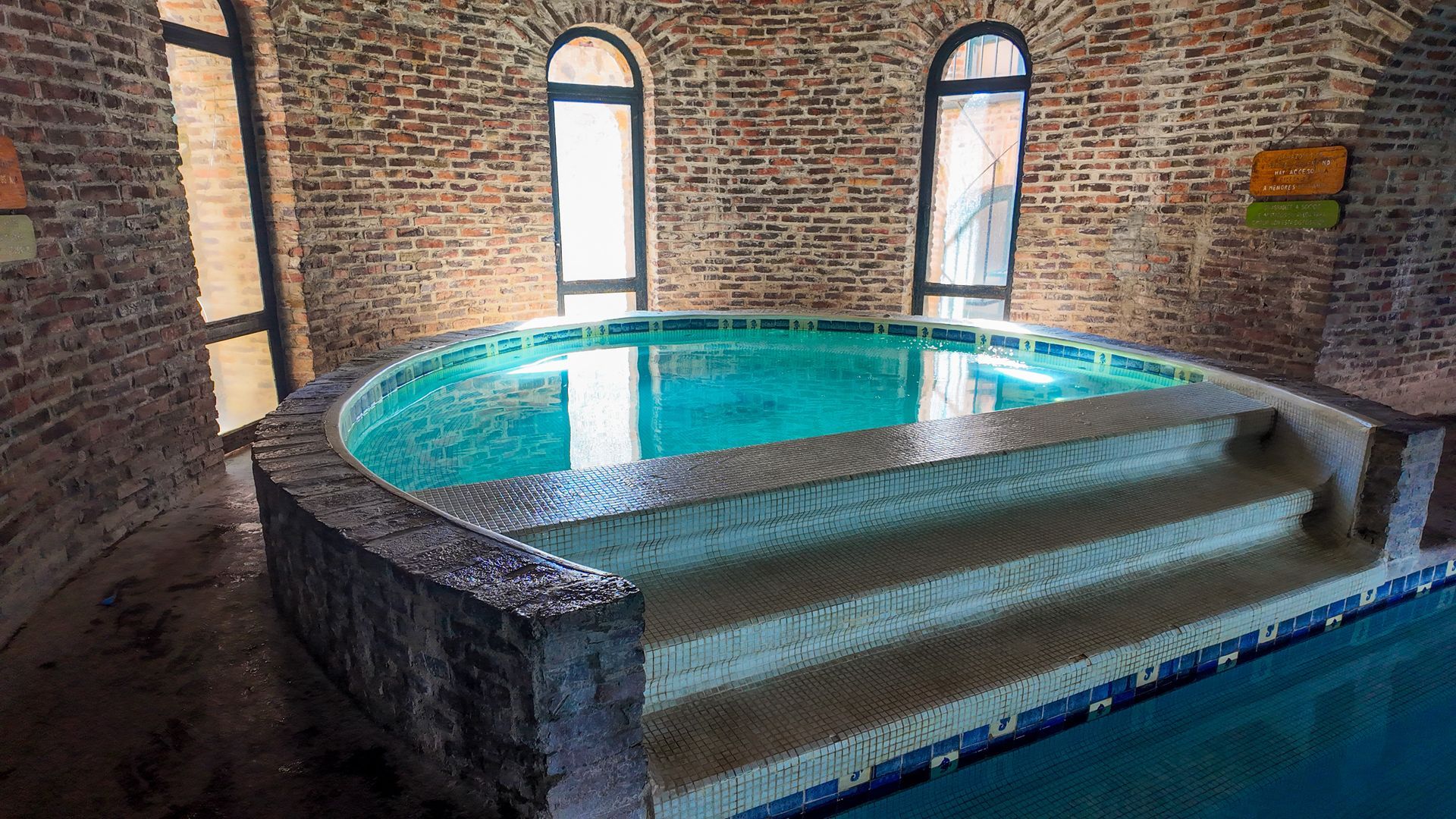 Piscina redonda cubierta con escalones, agua turquesa y paredes de ladrillo con altas ventanas arqueadas.