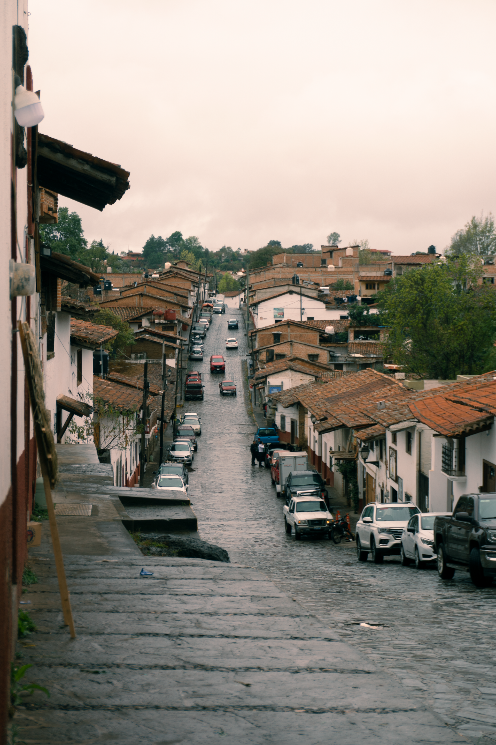 Calle empinada y empinada, bordeada de casas blancas y coches aparcados bajo un cielo nublado.
