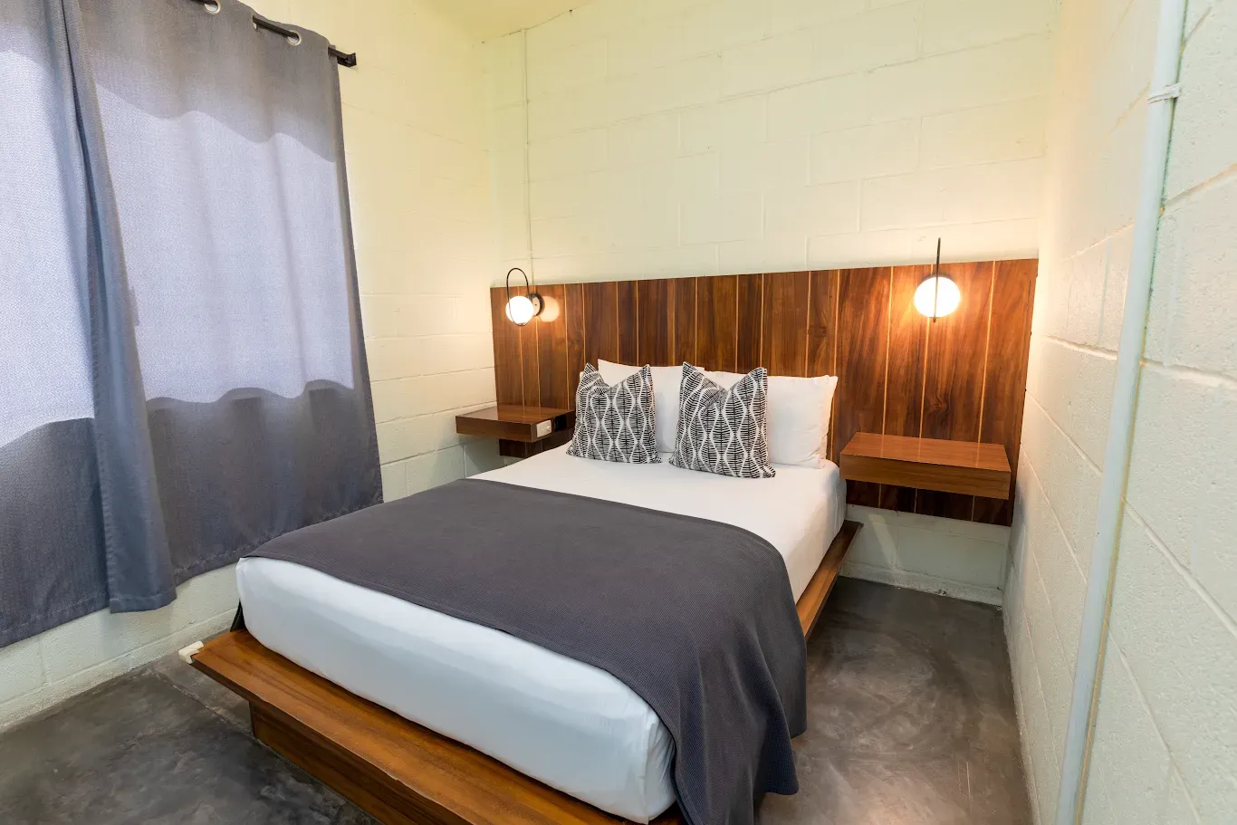 Cama bien hecha en un dormitorio sencillo con cabecero de madera, ropa de cama gris, lámparas de noche y una ventana con cortinas.