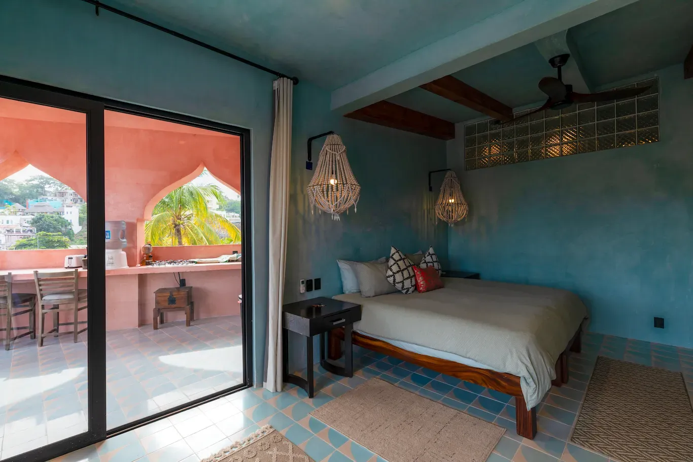 Habitación color turquesa con cama, decoración tejida y puertas corredizas que dan a un patio con vegetación tropical.