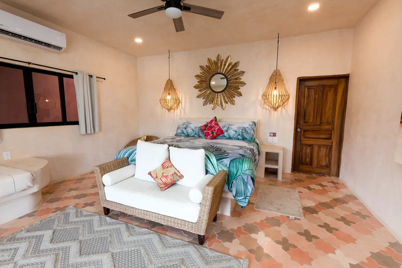 Acogedora habitación con dos camas individuales, sillas de mimbre, suelo de baldosas estampadas, ventilador de techo y puerta de madera rústica.
