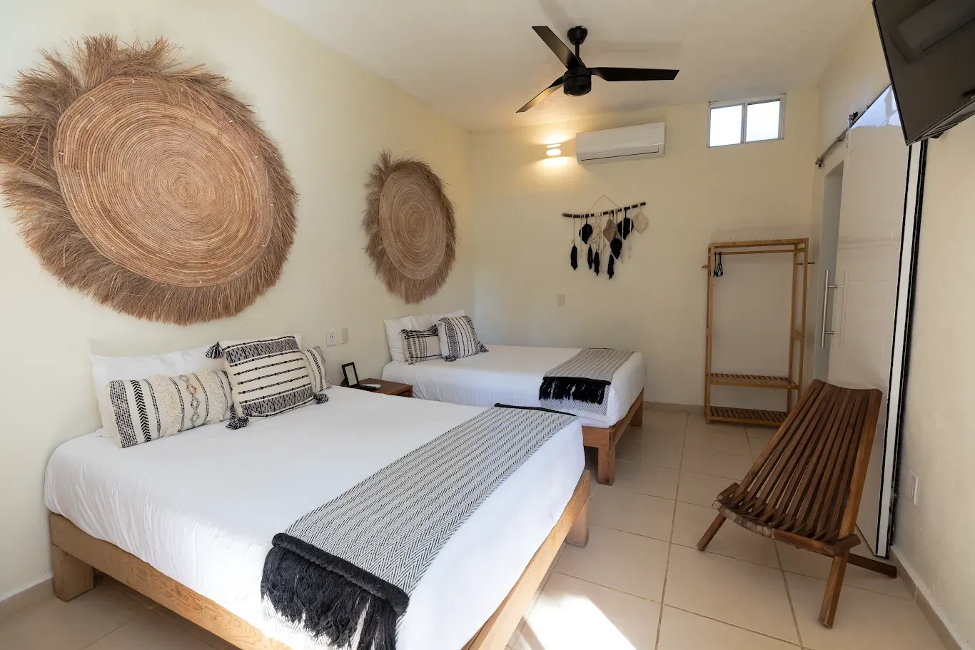 Habitación luminosa con dos camas blancas, decoración de pared tejida, ventilador de techo y silla hamaca cerca de una ventana.