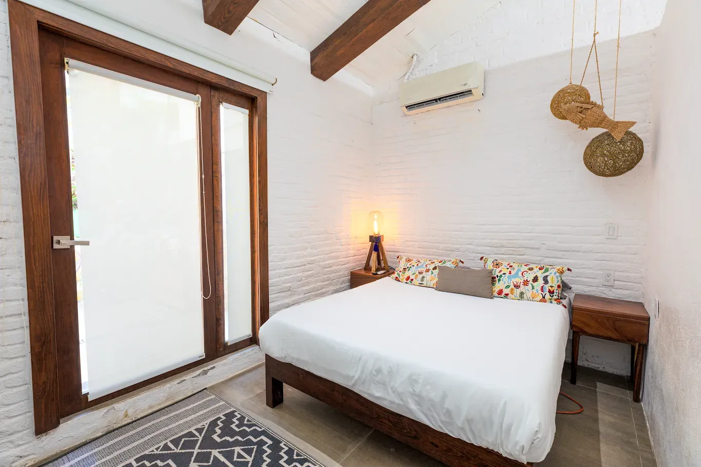 Habitación luminosa con cama, pared de ladrillo blanco, vigas de madera y puertas de cristal que dan a un balcón.