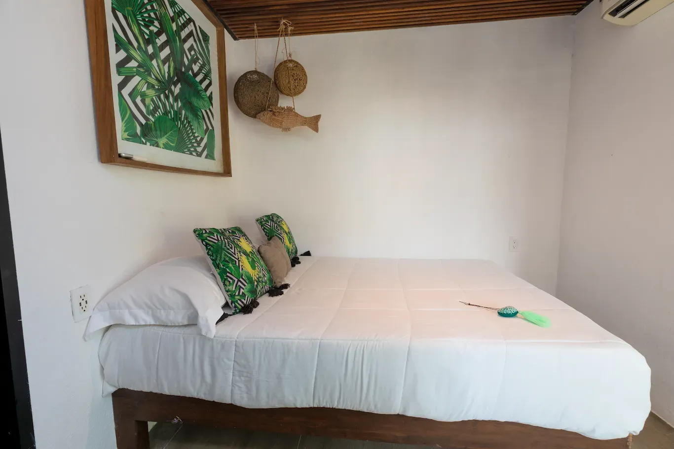 Habitación pequeña con cama blanca, almohadas verdes, decoración de pared tejida y techo de madera.