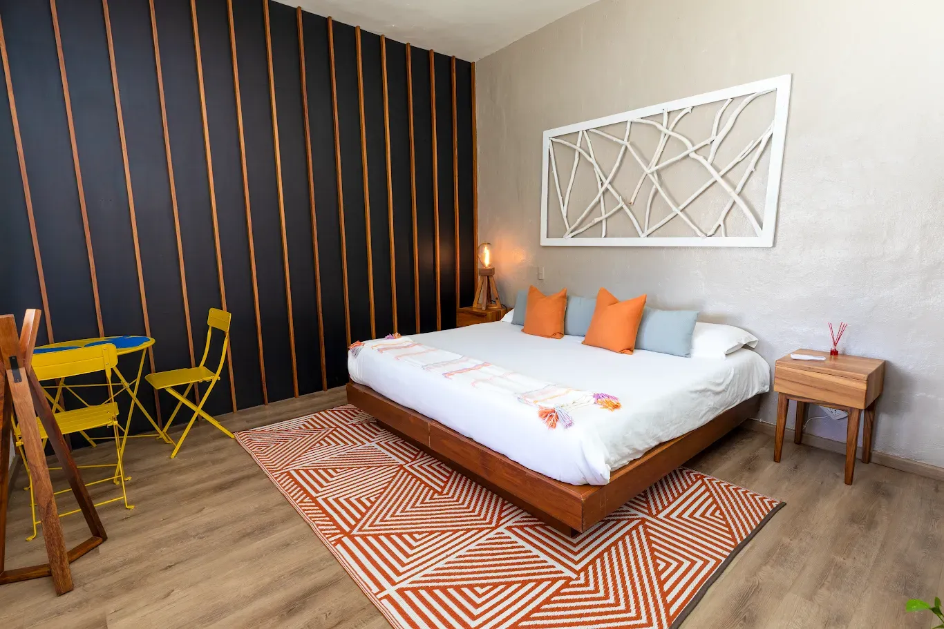Acogedora habitación con cama, detalles en naranja, alfombra estampada, pared decorativa oscura y pequeña zona de estar.