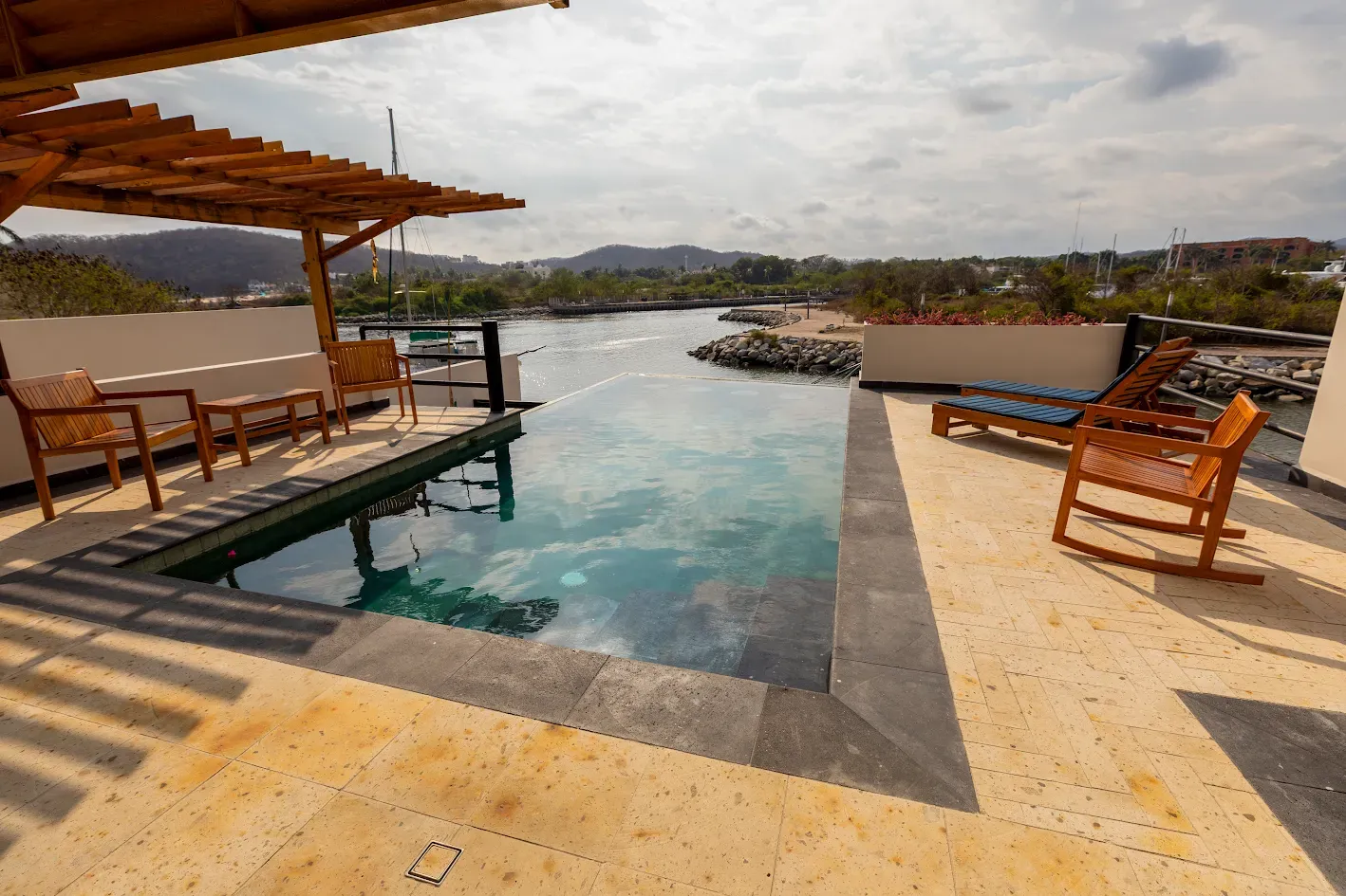 Piscina infinita junto a una terraza al borde del lago con sillas de madera y una pérgola, con vistas a las montañas bajo un cielo nublado.