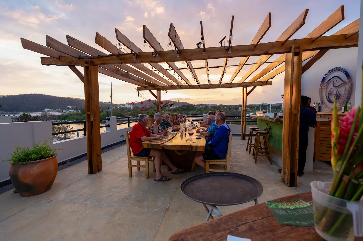 Gente cenando en una mesa al aire libre en una azotea, bajo una pérgola de madera, al atardecer, con vistas a la ciudad.
