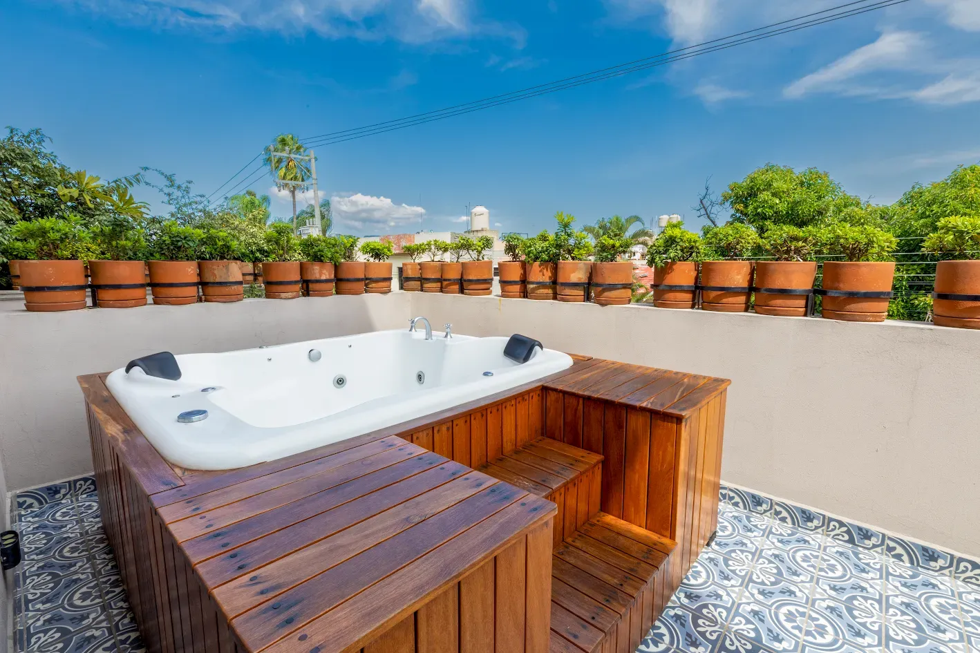 Bañera de hidromasaje al aire libre en una terraza embaldosada con bancos de madera y plantas en macetas bajo un cielo azul.