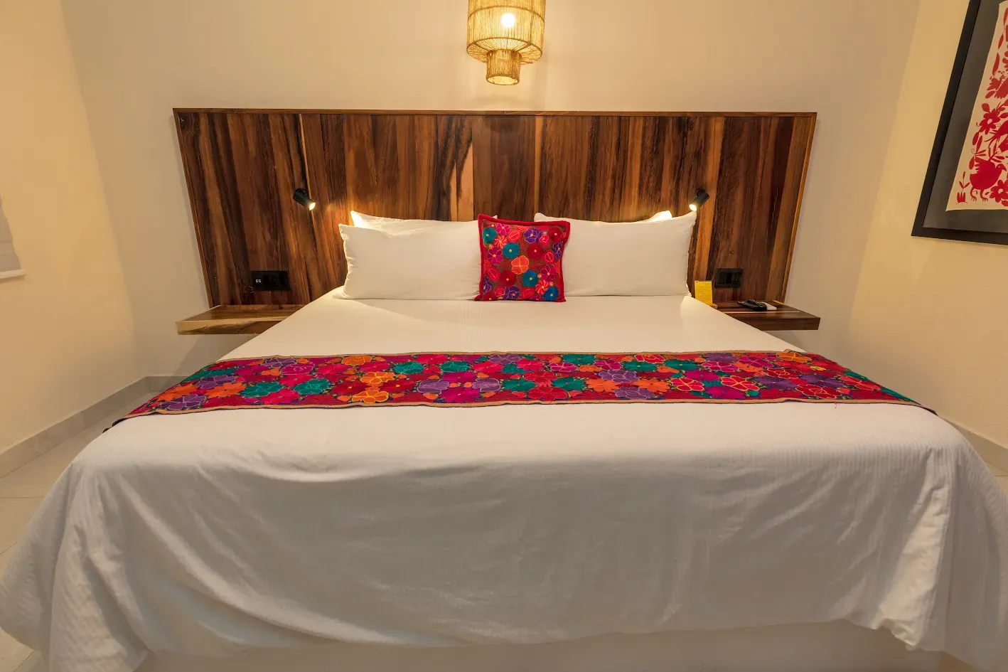 Cama bien hecha con sábanas blancas y un colorido camino de mesa y almohadas rojas y azules en una habitación de hotel con iluminación cálida.